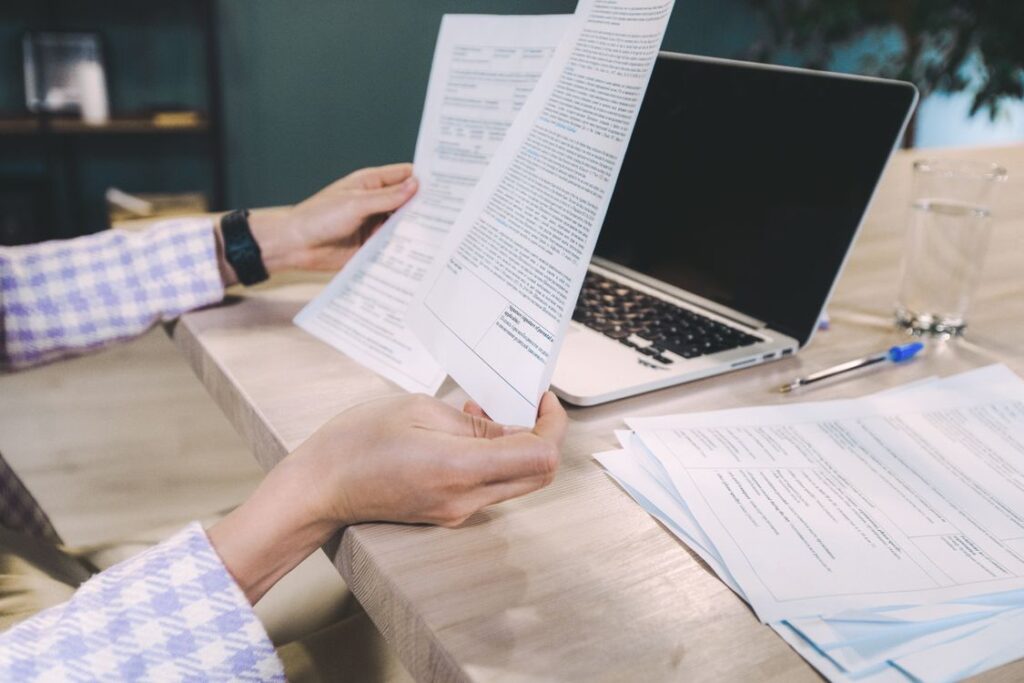 Hands review printed documents beside a laptop.