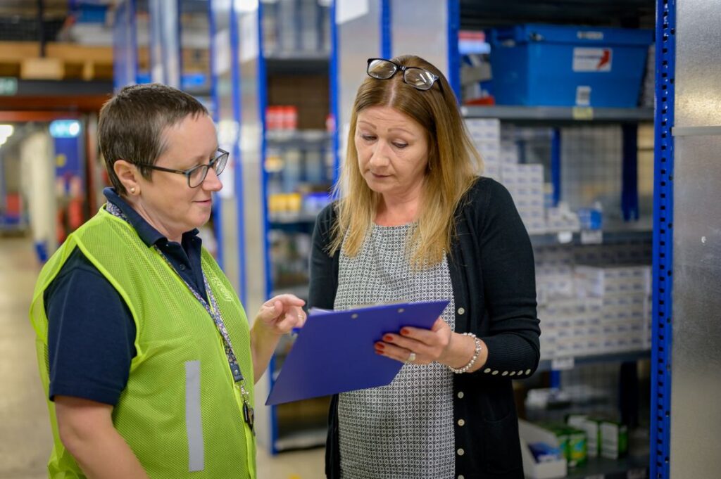 Two warehouse colleagues check a clipboard beside storage shelves.