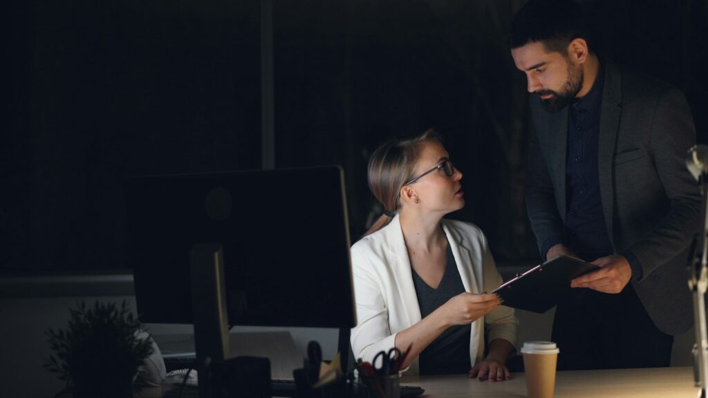 Two colleagues review a clipboard beside a computer during an evening shift.