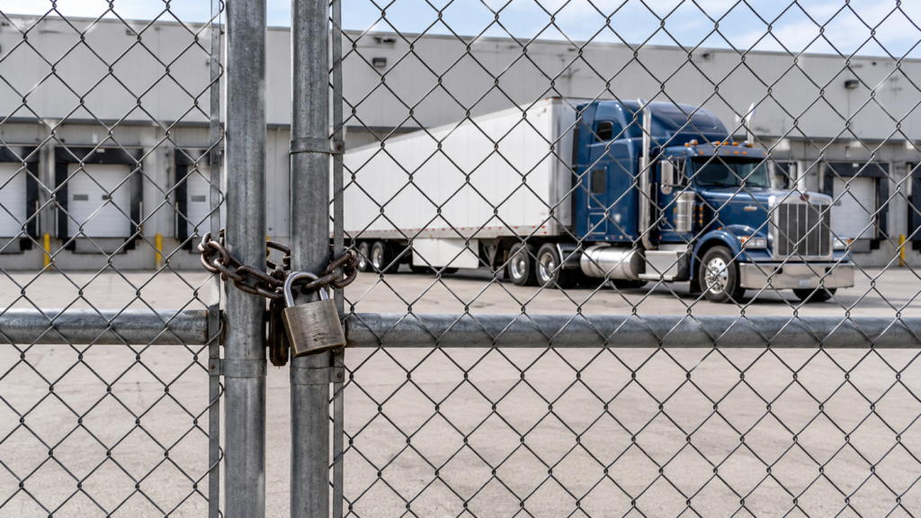 Illustrative scene of a locked freight-yard gate with an American long-hood semi-truck and trailer behind it.