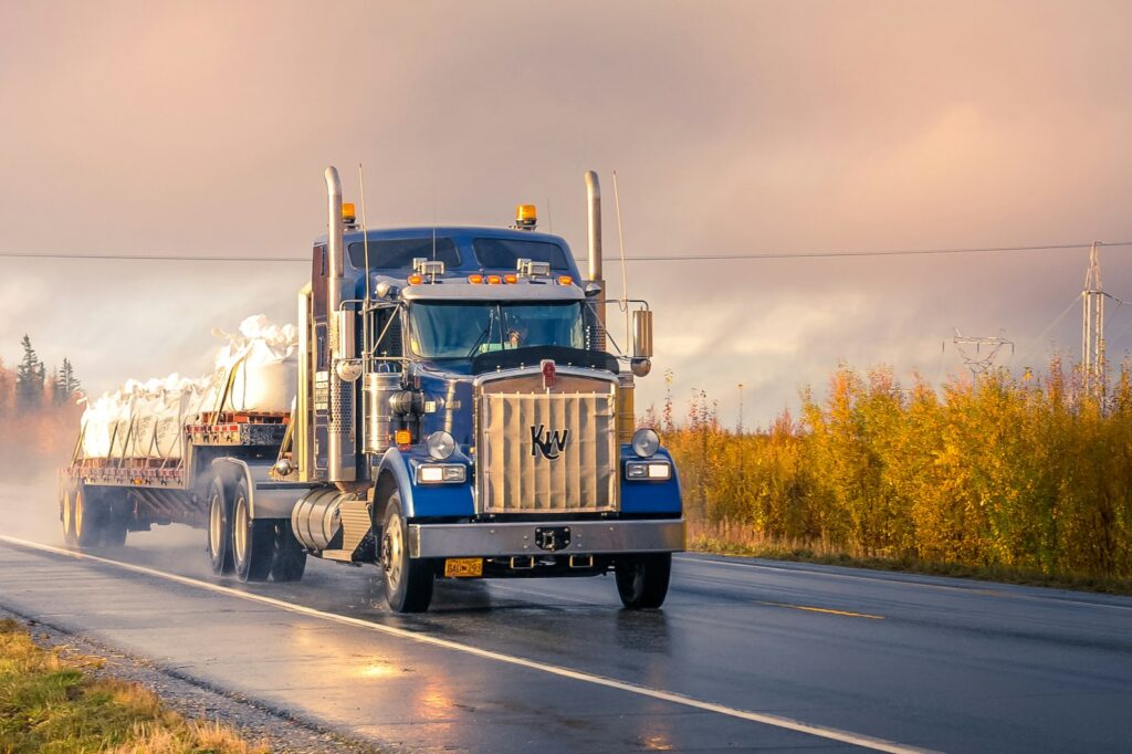Blue conventional semi-truck hauling a flatbed trailer on an Alaska highway.