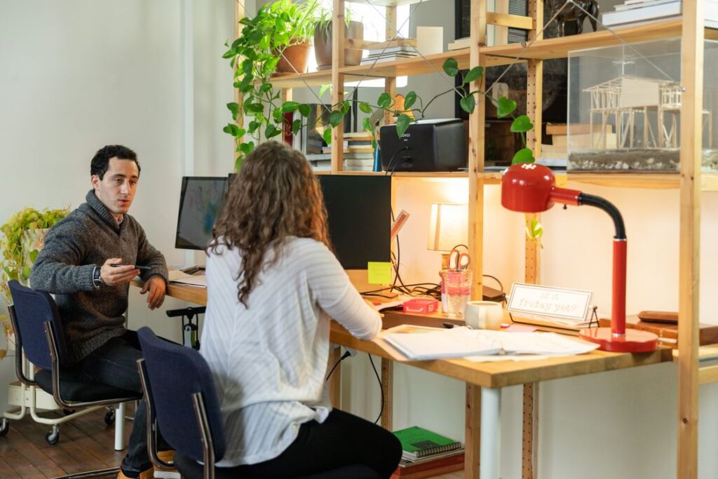 Two colleagues discuss work beside computer monitors and an open notebook.